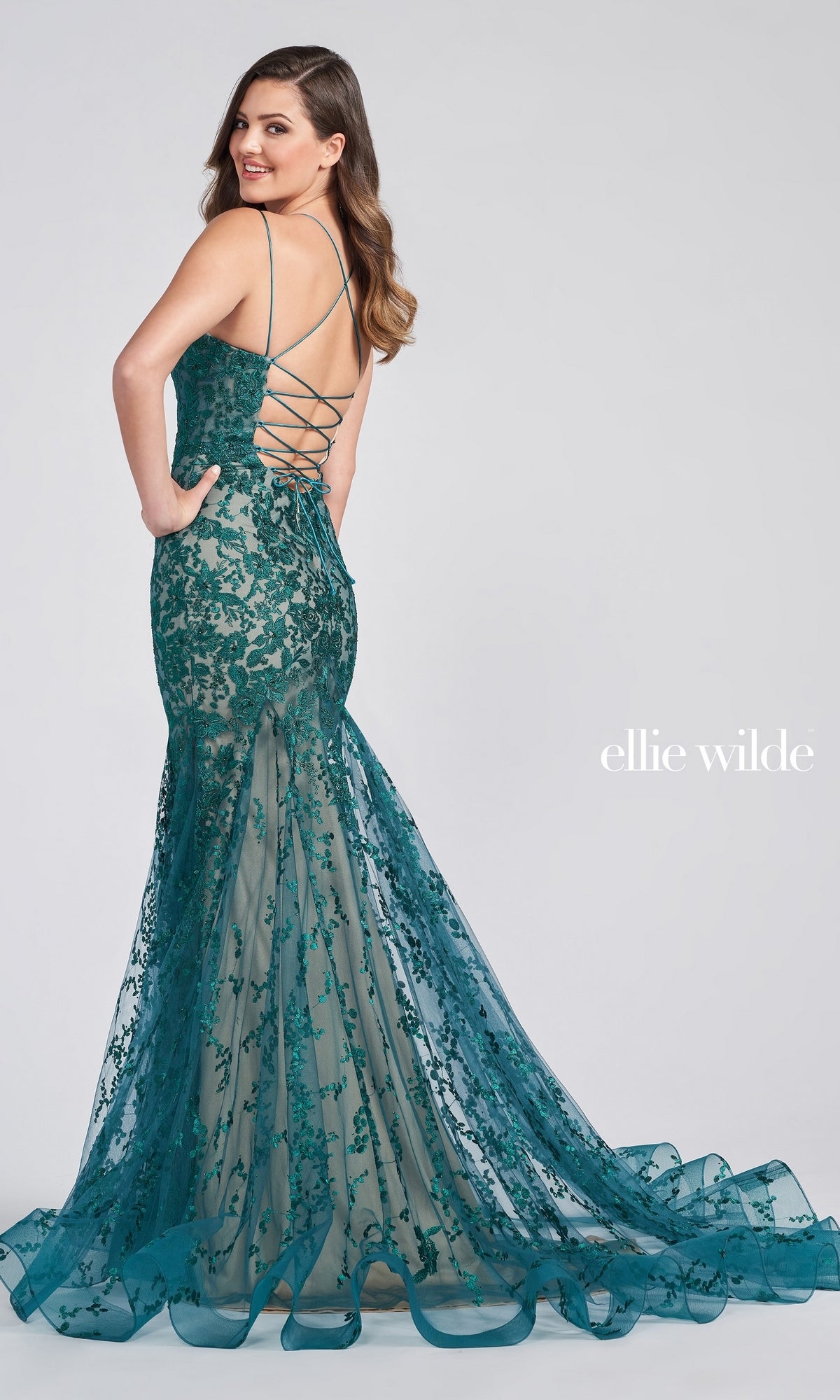 A woman wears a Long Lace Mermaid Prom Dress with a strappy back by Ellie Wilde, posing gracefully against a plain light background.